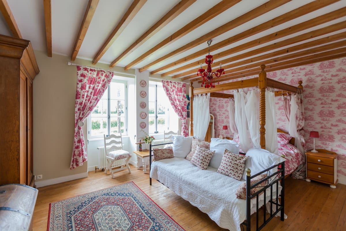 Ein Schlafzimmer mit einem Himmelbett, Holzbalkendecke und einem großen Fenster mit Vorhängen.