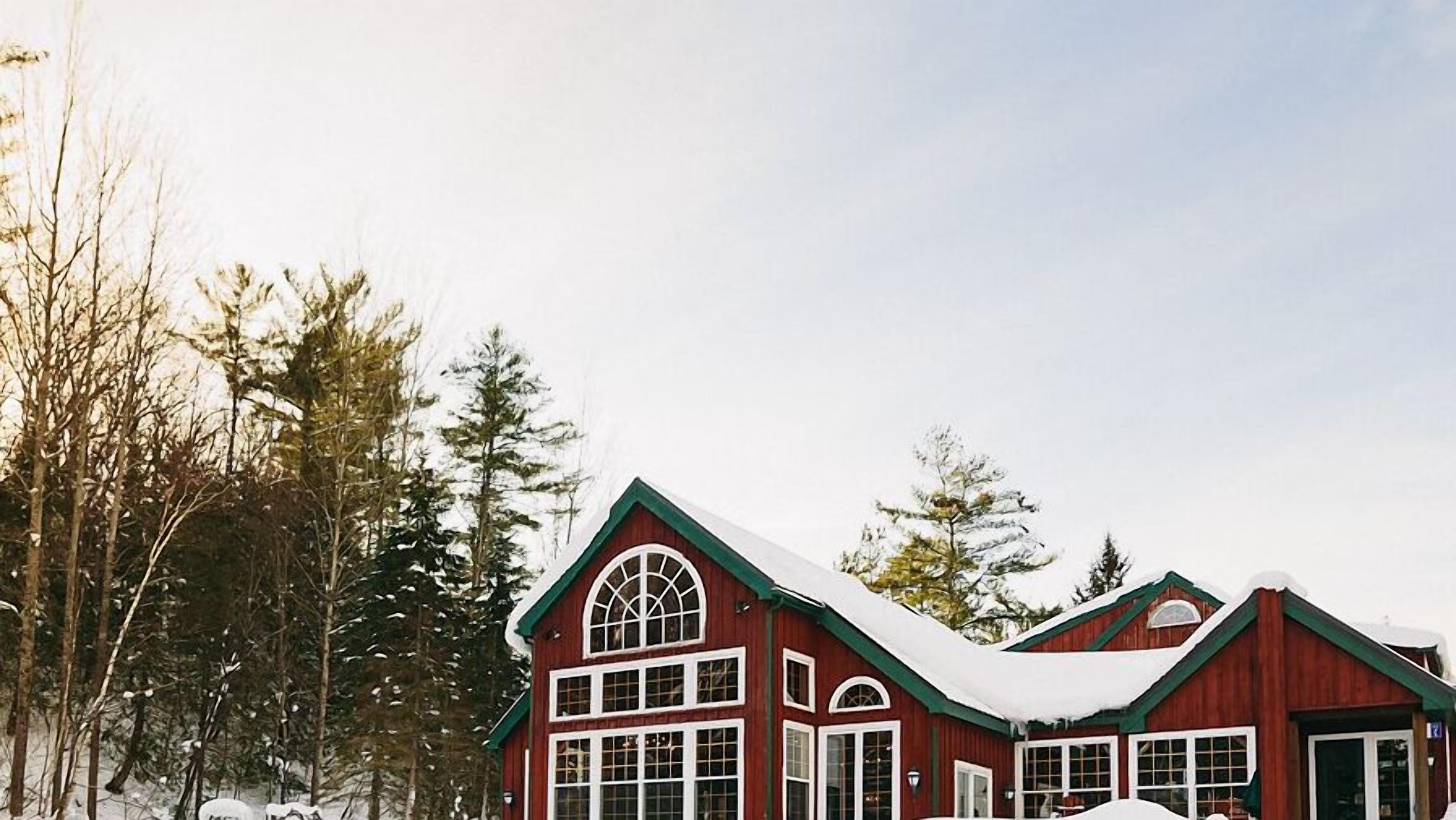A red wooden building with multiple windows and a snow-covered roof, surrounded by trees.