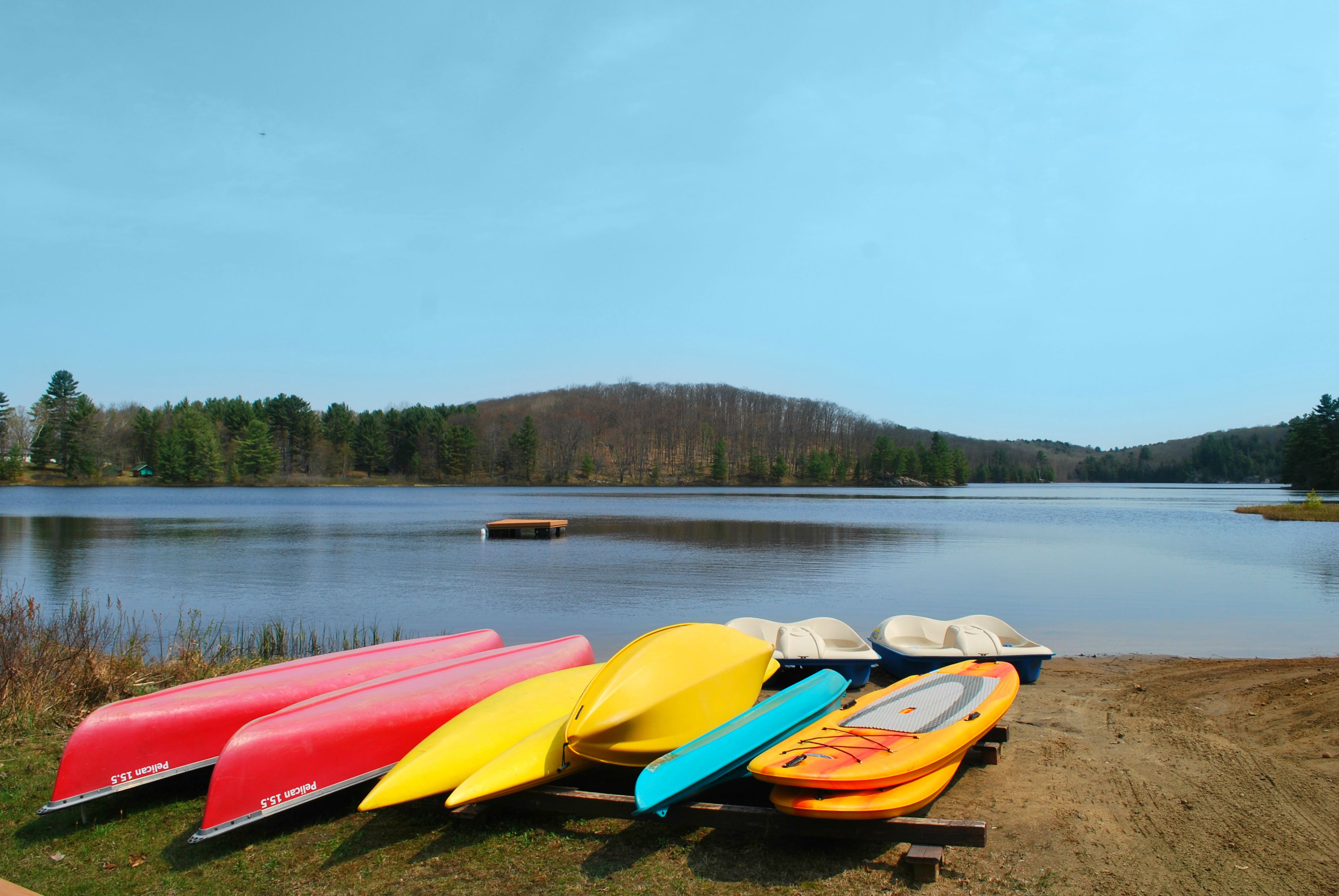 A row of colorful kayaks lined up on a wooden dock by a calm lake with a forested hill in the background.