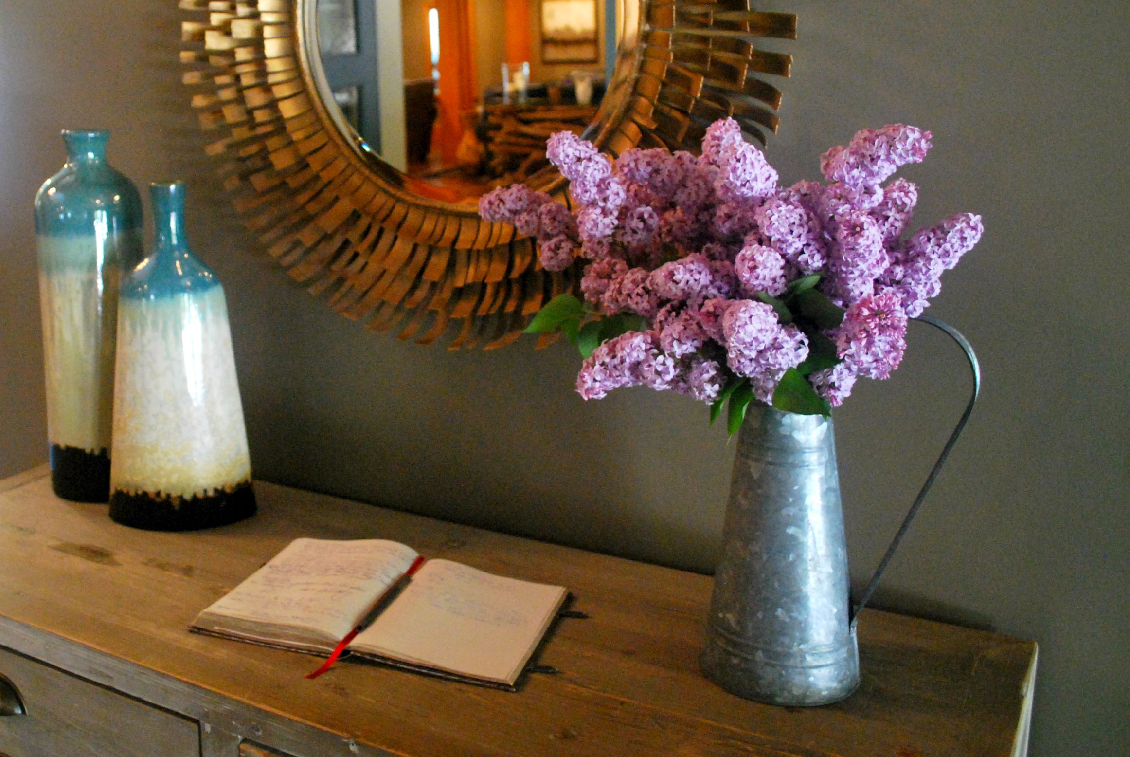 A wooden dresser with a mirror, a vase of purple flowers, and two bottles.