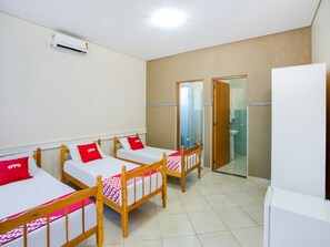 Une chambre d’hôtel avec quatre lits simples, chacun doté d’un cadre en bois et d’une literie rouge et blanche. On aperçoit un climatiseur blanc fixé au mur, un réfrigérateur blanc et une salle de bain à travers une porte ouverte.