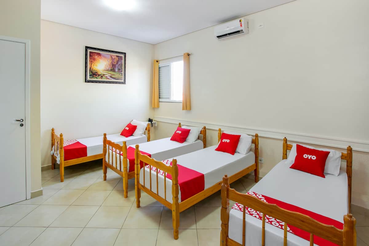 Ein Hotelzimmer mit vier Einzelbetten, jeweils mit Holzrahmen und Bettwäsche in Rot und Weiß. An der Wand hängt ein Gemälde, ein Fenster mit Vorhängen und eine Klimaanlage sind ebenfalls vorhanden.