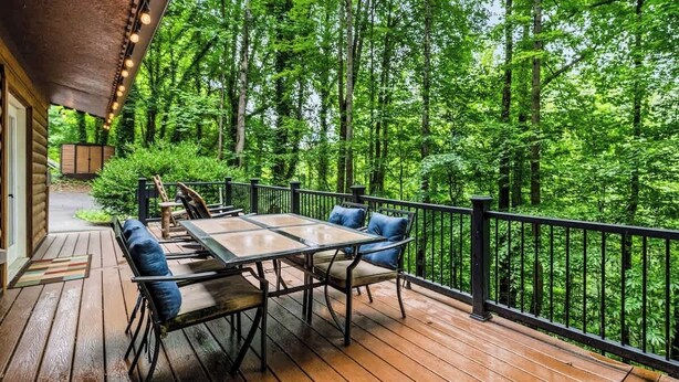 A wooden deck with a table and chairs, surrounded by a black railing, overlooking a forested area.