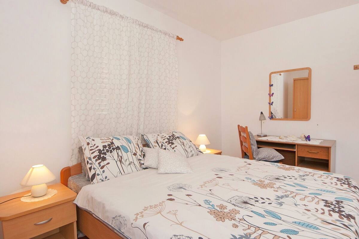 A bedroom with a bed, bedside table, lamp, desk, chair, and a mirror.