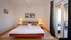 A bedroom with a large bed, two bedside tables with lamps, a painting on the wall, and a sliding glass door to the right. - Apartments Mara (Supetar)