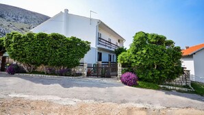 A two-story white house with a balcony, surrounded by greenery and a stone wall. - Apartments Jelica (Pag)