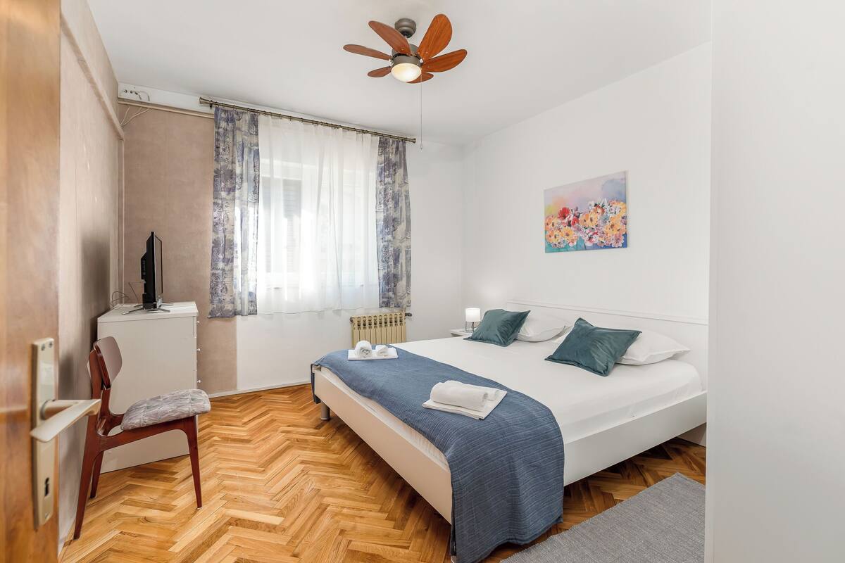 A bedroom with a bed, a chair, a desk, a radiator, and a ceiling fan.