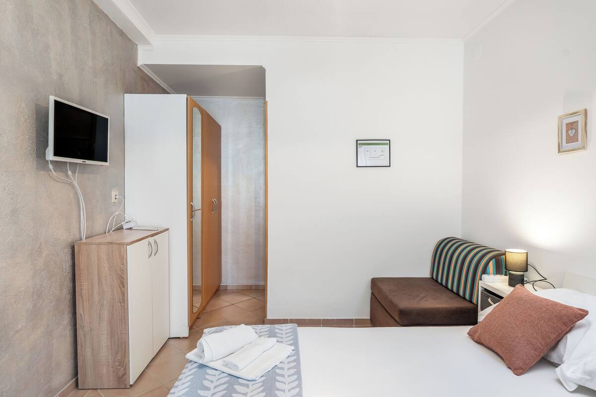 A modern hotel room with a bed, a bedside table with a lamp, a TV mounted on the wall, a wooden cabinet, and a door leading to another room.