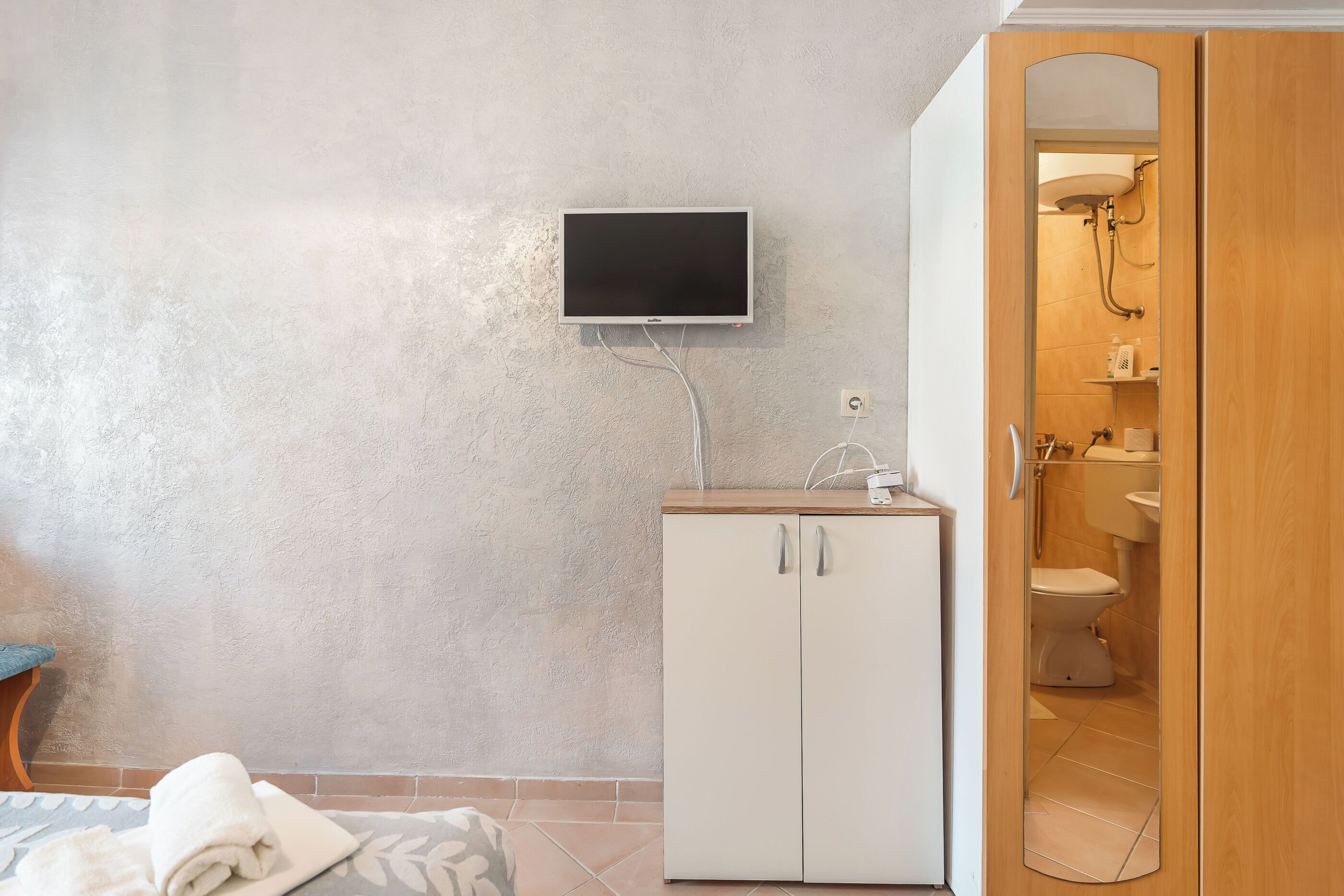 A room with a television mounted on the wall, a wardrobe, and a bathroom visible through an open door.