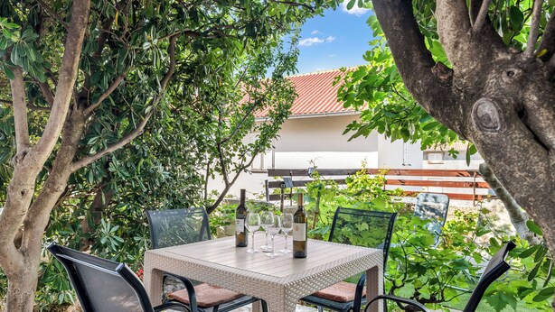 An outdoor patio with a table set for four, surrounded by chairs, and a view of a building with a red roof. - Apartments Jelica (Pag)