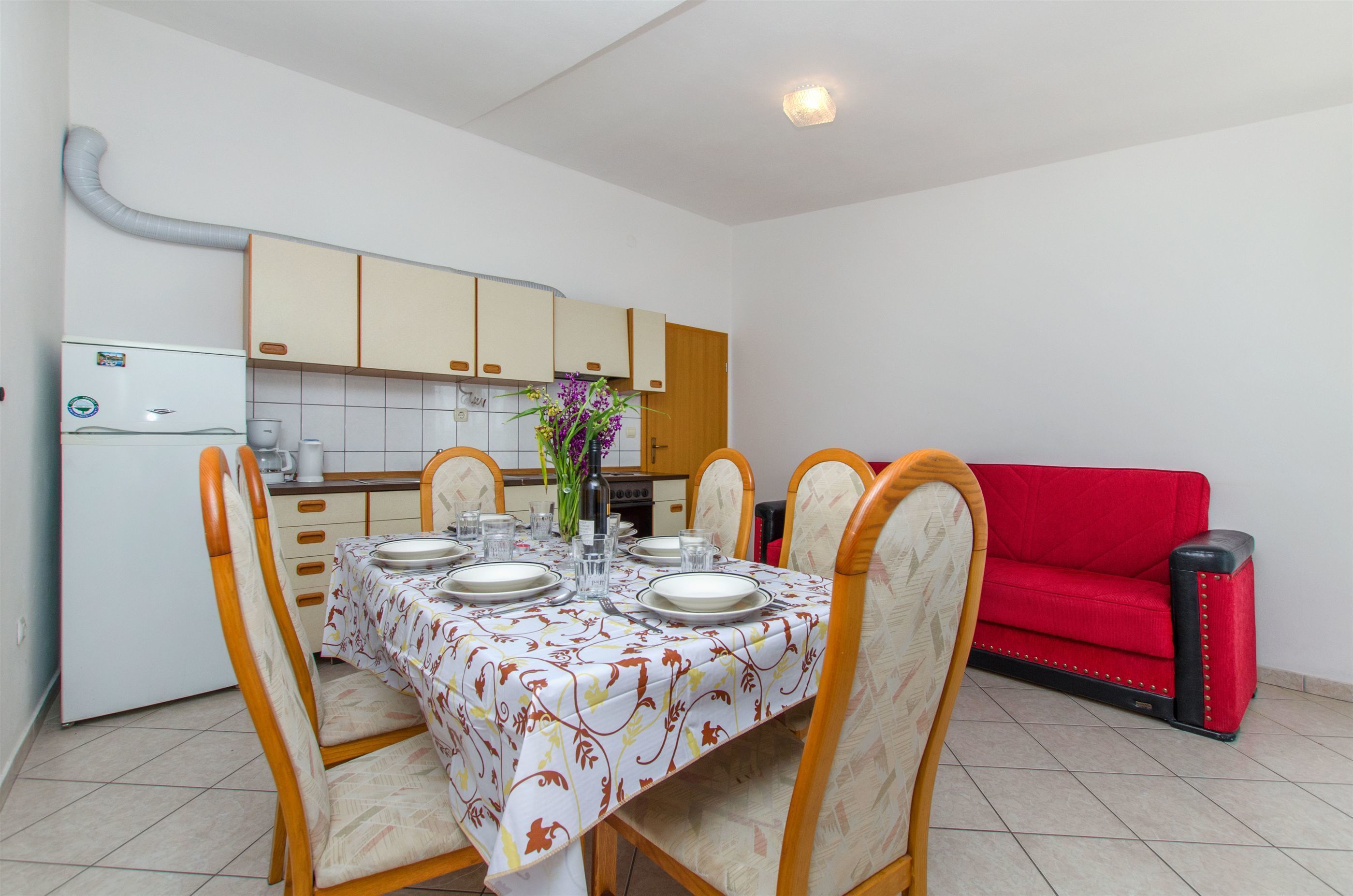 Un coin repas avec une table dressée pour quatre personnes, entourée de chaises. Un canapé rouge est visible au fond.