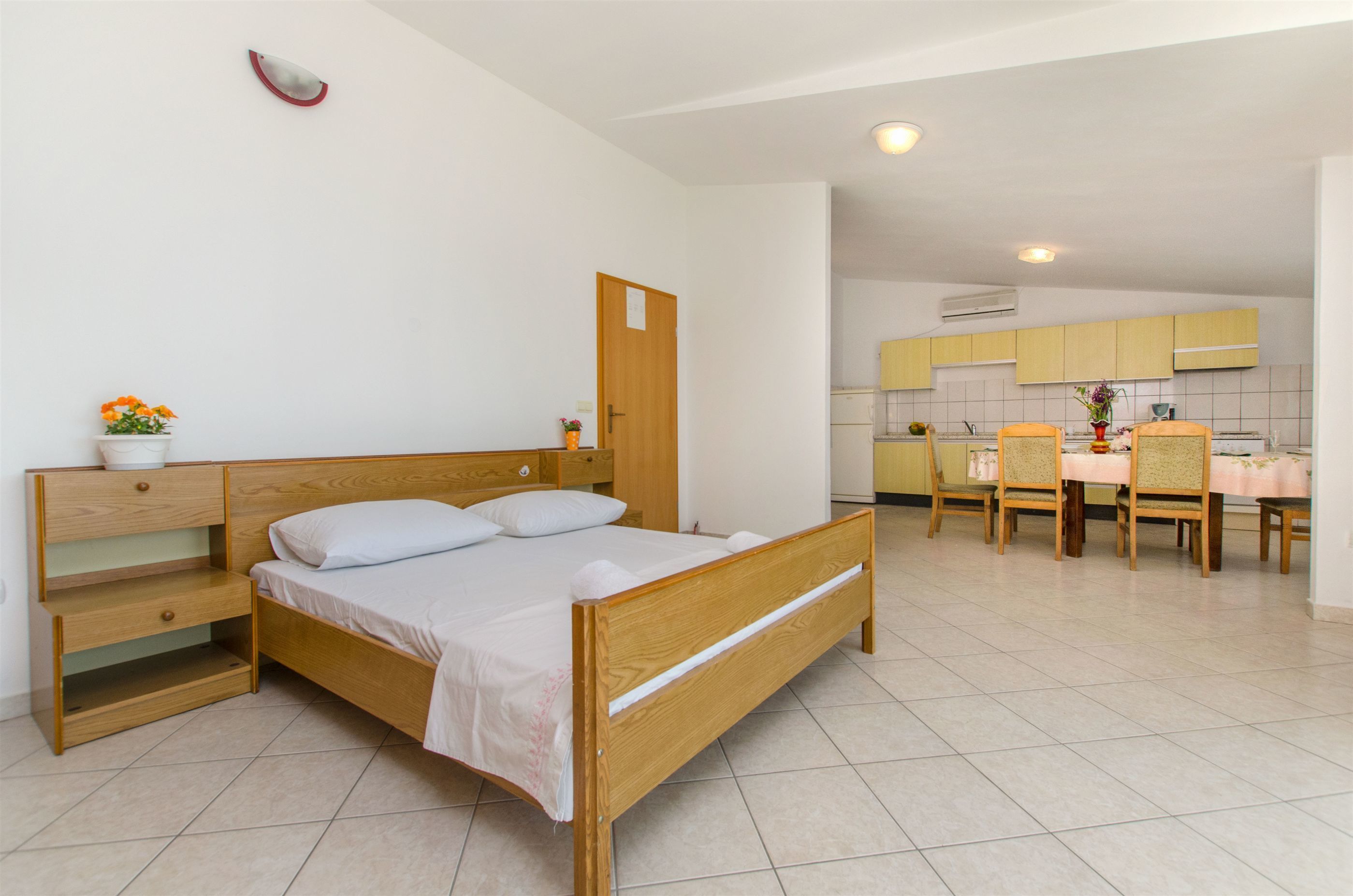 Ein Schlafzimmer mit einem hölzernen Bett, einem Nachttisch, einem kleinen Tisch mit Blumenvase und Blick in eine Küche mit gelben Schränken und einem Essbereich.