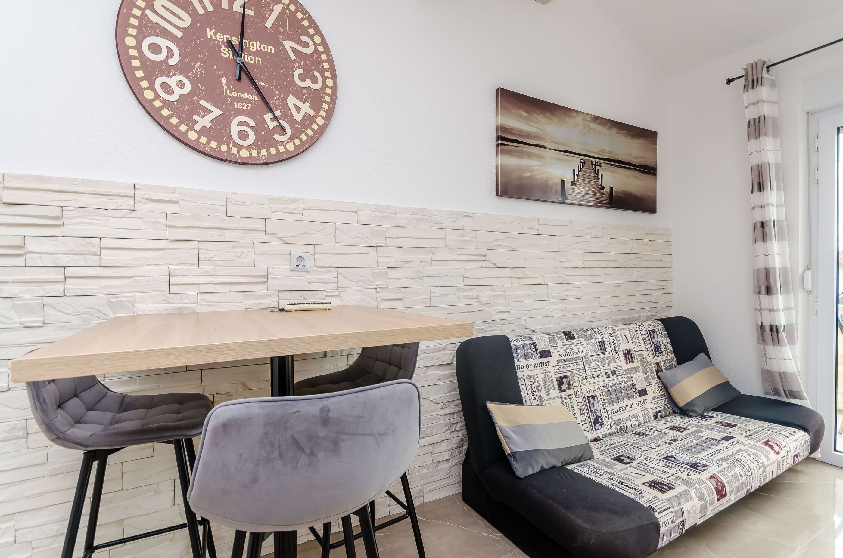 A modern living room with a wooden table, two grey chairs, a black sofa with patterned cushions, a large wall clock, and a painting of a sunset.