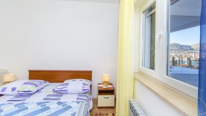 A bedroom with a bed, bedside table, lamp, and a window with a view of a cityscape. - Apartment Marijana (Trogir)