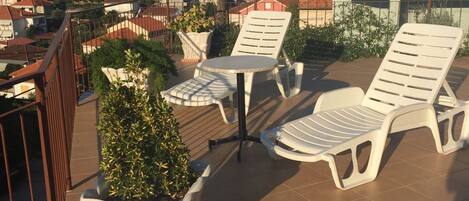 Una terraza con sillas de plástico blanco y una mesa, rodeada de plantas en macetas.