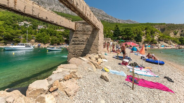 A coastal scene with a rocky beach, clear turquoise water, boats, and a stone bridge. - Apartments Stjepan (Lokva Rogoznica)