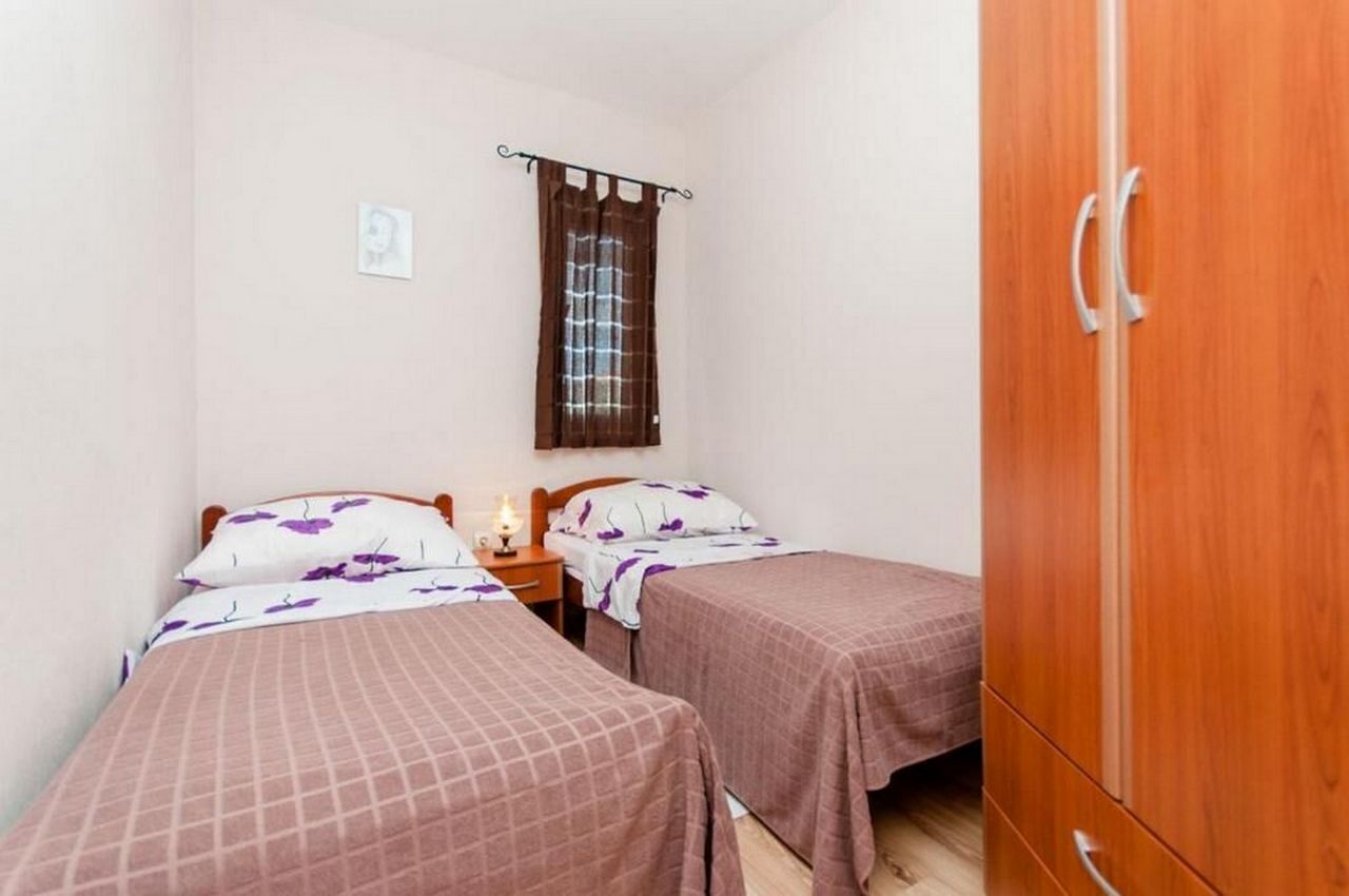 Quarto pequeno com duas camas de solteiro, um guarda-roupa de madeira, uma mesa de cabeceira com luminária e uma janela com cortinas.