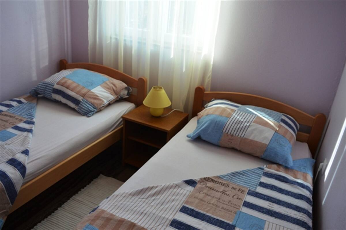 Habitación doble con dos camas individuales, cada una con un almohadón a rayas y un cabecero de madera. Entre las camas, una mesita de noche de madera con una lámpara amarilla.
