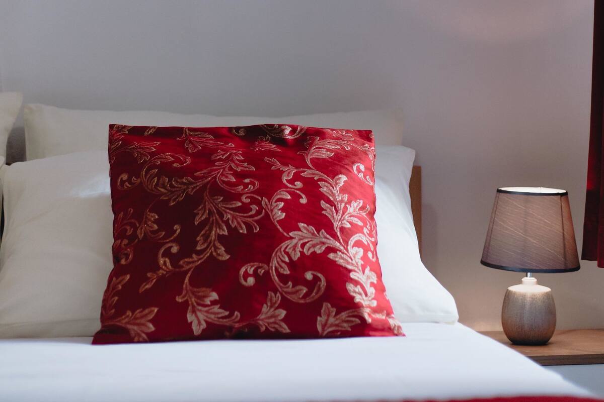 Una cama con almohadas blancas y rojas, una almohada con estampado floral rojo, una mesita de noche con lámpara y adornos de mariposas en la pared.