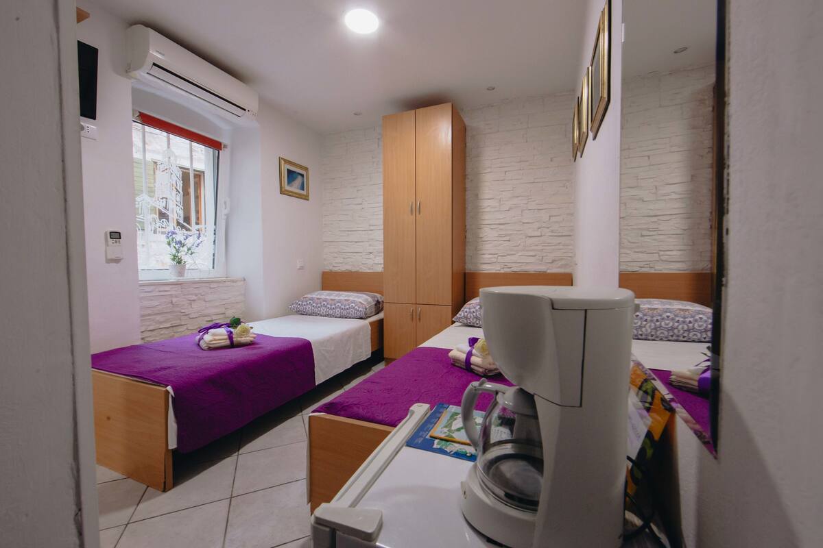 Un dormitorio pequeño y bien iluminado con dos camas, un armario, un equipo de aire acondicionado y una ventana con vistas.