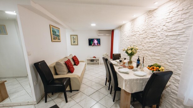 A dining area with a white table set for four, surrounded by black chairs. A beige sofa with red cushions is against the wall, and a flat-screen TV is mounted above it. - Apartment and Room Old Town (Split)