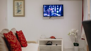 A living room with a television mounted on the wall, a white sofa with red cushions, a small white side table with a flower, and a potted orchid. - Apartment and Room Old Town (Split)