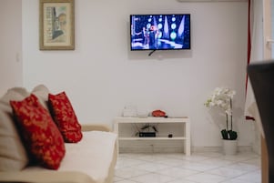Egy nappali, melyben a falra van szerelve egy televízió, egy fehér kanapé piros párnákkal, egy kis, fehér dohányasztal, rajta egy virággal, és egy virágozó orchidea található.