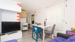 Una sala moderna con un televisor de pantalla plana, un comedor con mesa blanca y sillas azules, y una cocina con gabinetes blancos y un electrodoméstico amarillo.