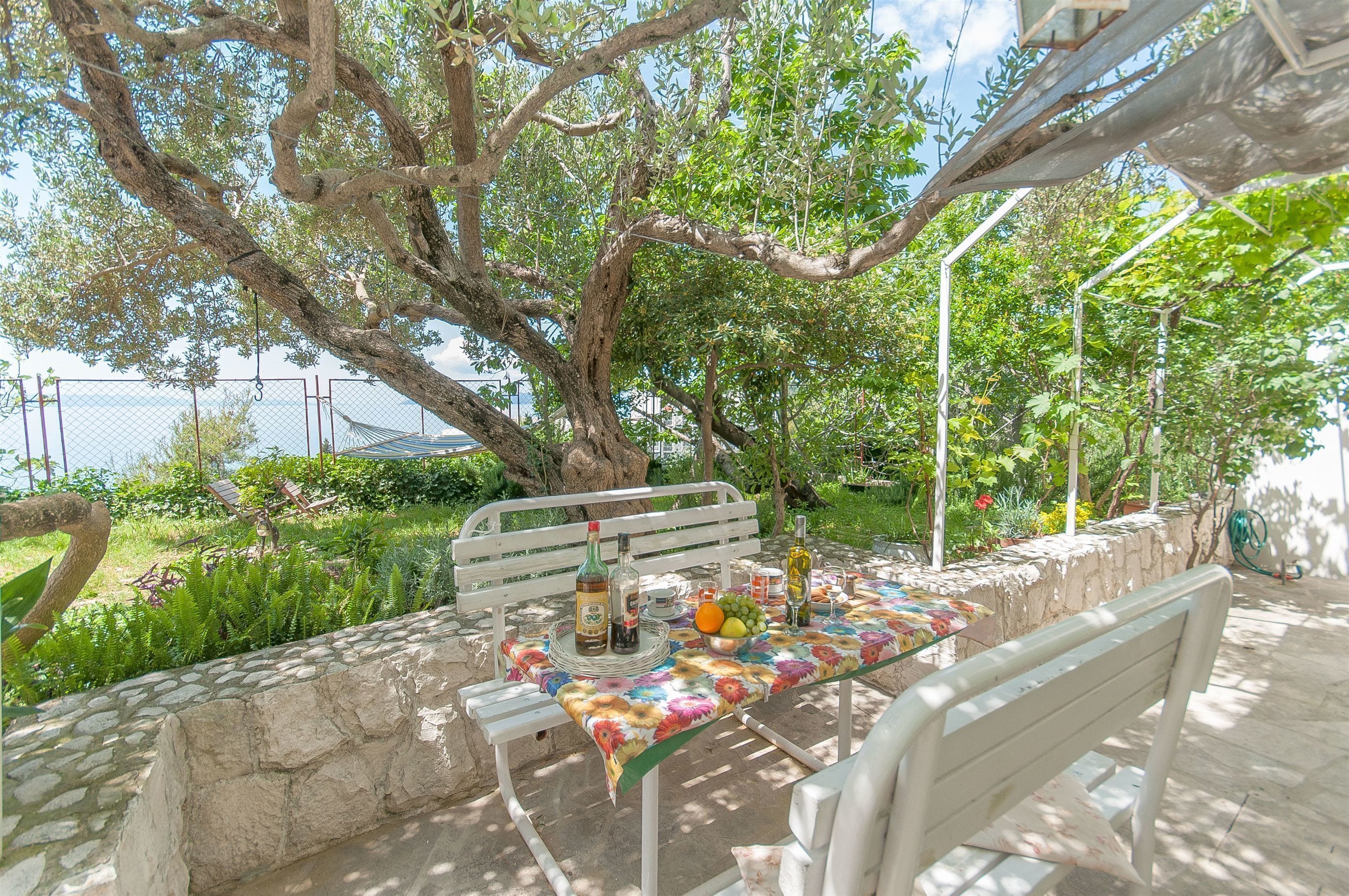 An outdoor seating area with a table set for two, surrounded by greenery and a stone wall.