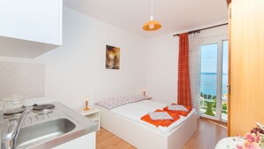 Habitación compacta con kitchenette, cama con ropa de cama naranja, un cuadro en la pared y vista al mar a través de una puerta corrediza de vidrio.