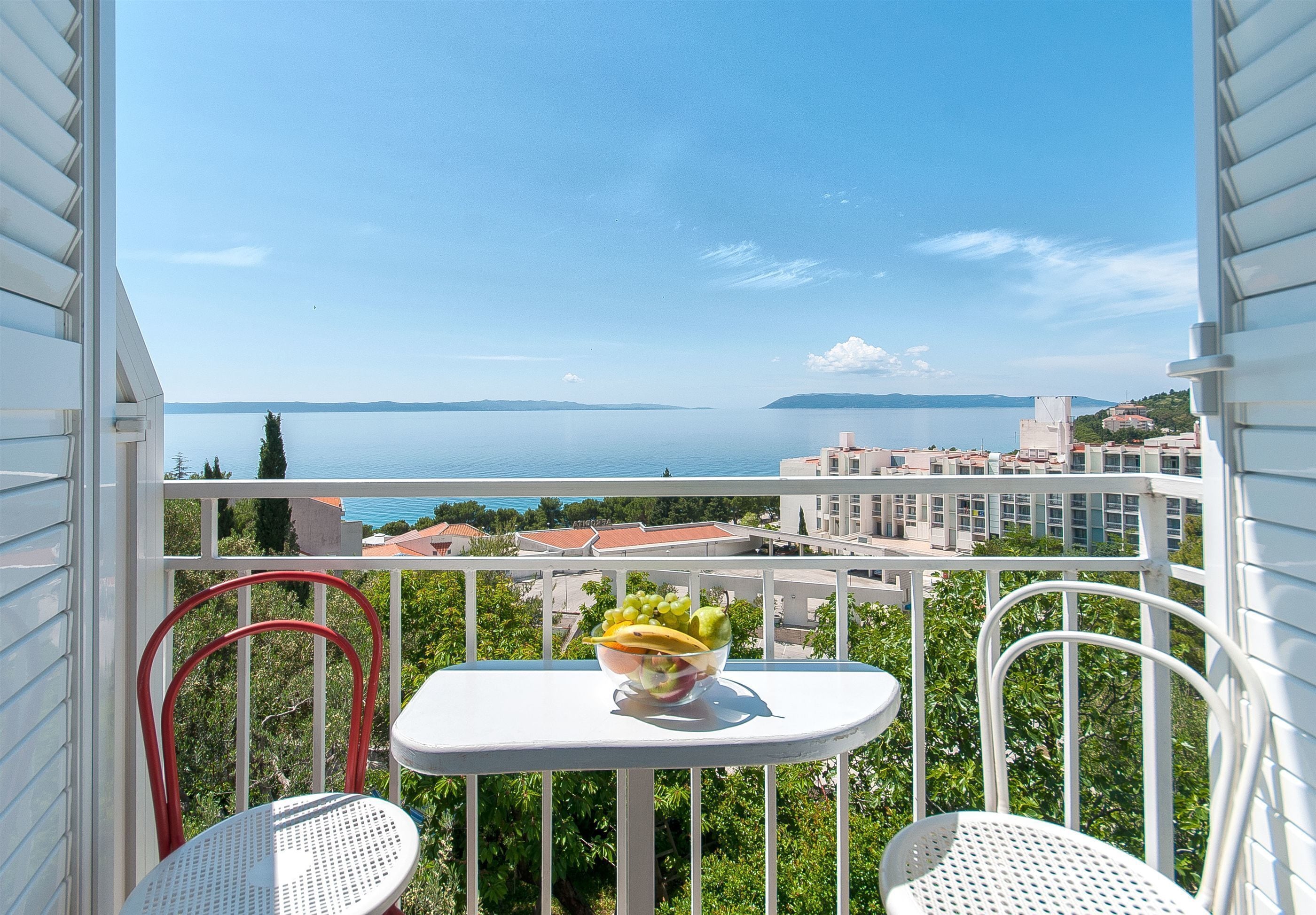 A balcony with a table and two chairs, a bowl of fruit, and a view of the sea and buildings.
