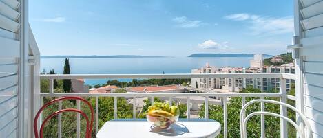 A balcony with a table and two chairs, a bowl of fruit, and a view of the sea and buildings.