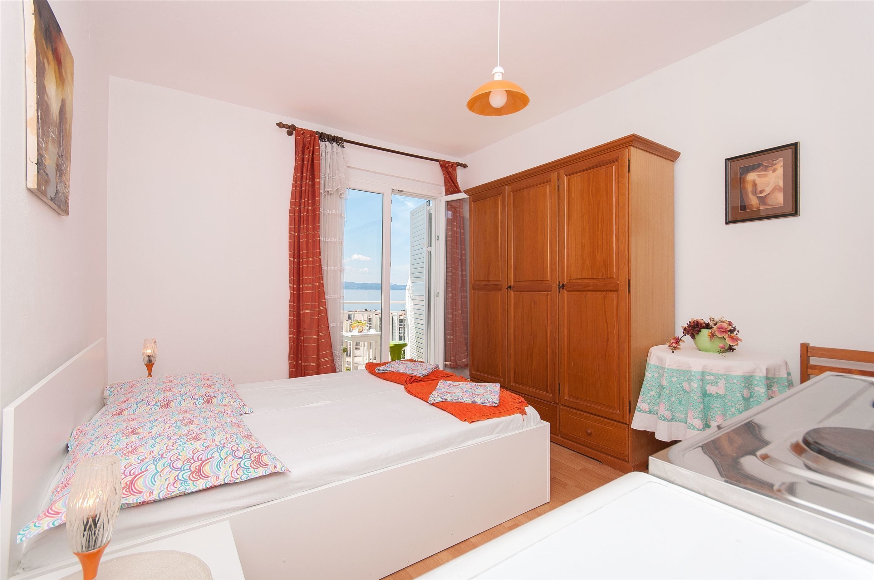 A bedroom with a bed, a wooden wardrobe, a small table with a vase, and a view through a window.