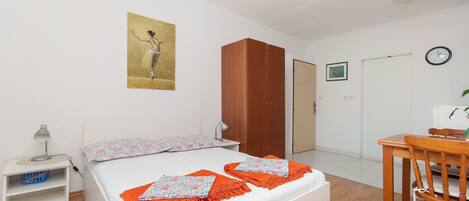 A bedroom with a bed, a wooden wardrobe, a desk with a chair, a wall clock, and a painting.