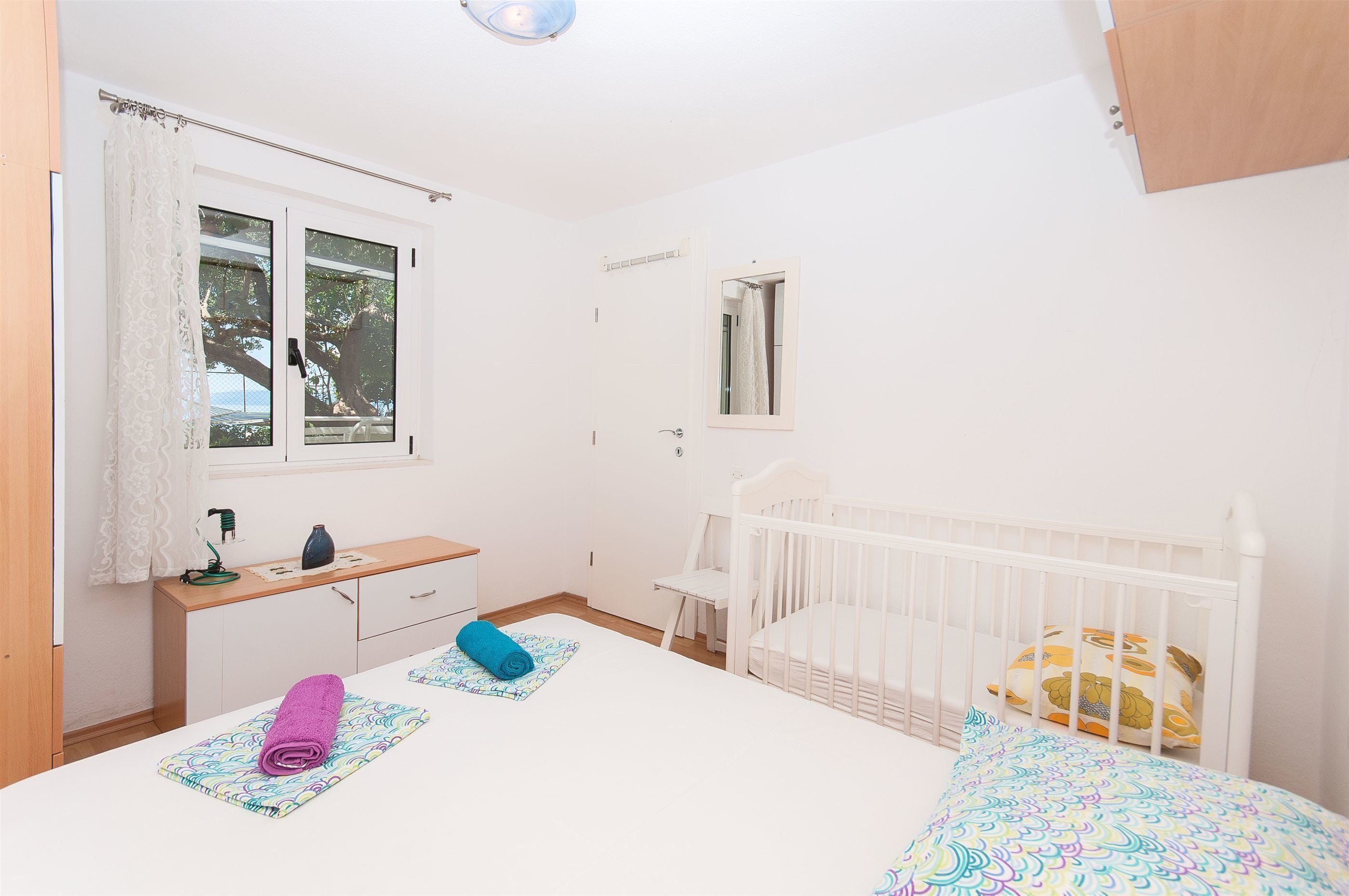 Une chambre avec un berceau, une fenêtre avec des rideaux, une table de chevet et un miroir.
