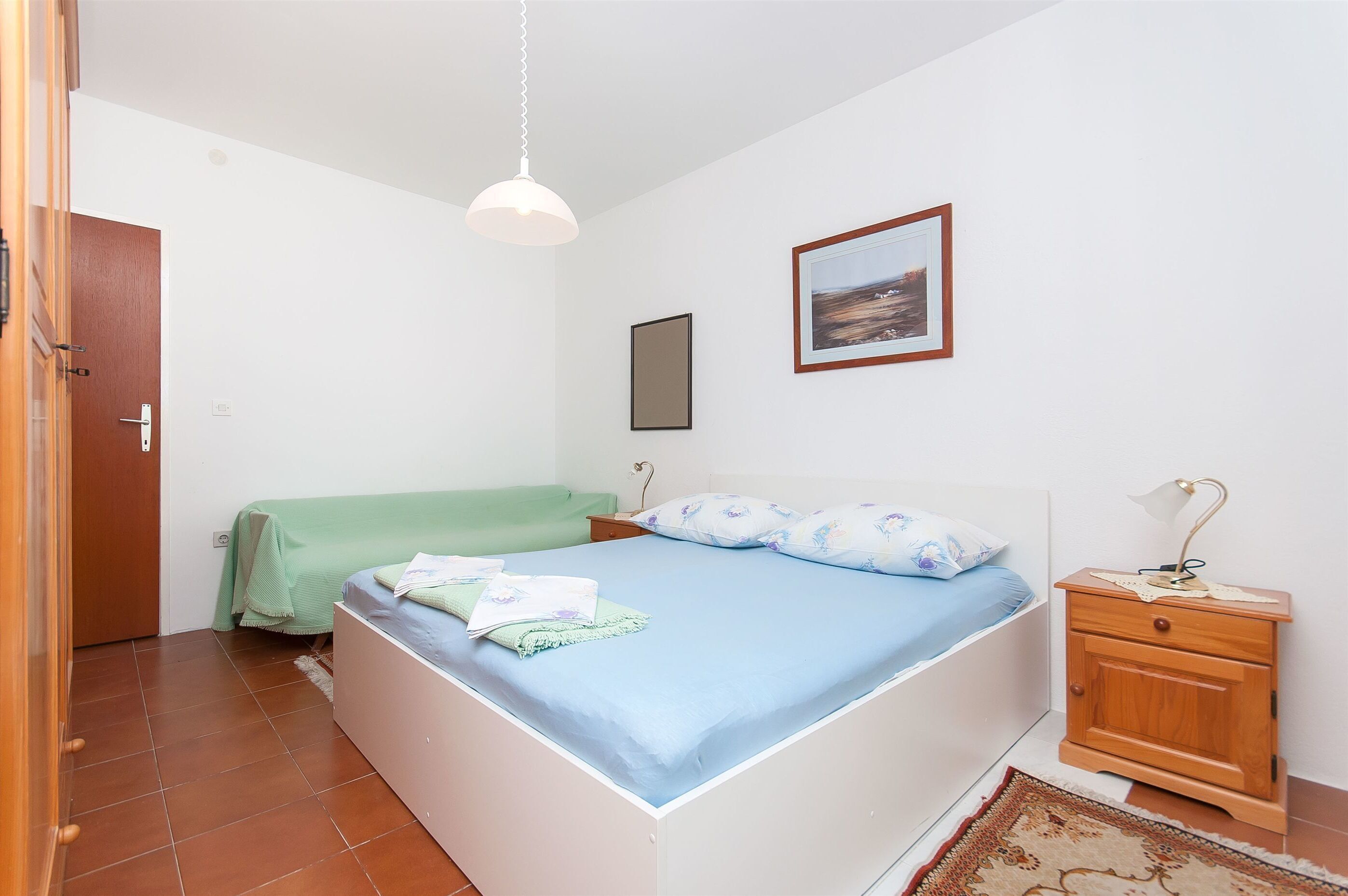 Un dormitorio con cama, mesita de noche, lámpara colgante, un cuadro enmarcado en la pared y una puerta.