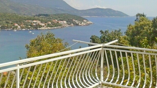 Un balcon doté d’une balustrade blanche, qui offre une vue sur un paysage côtier avec des collines et une étendue d’eau.
