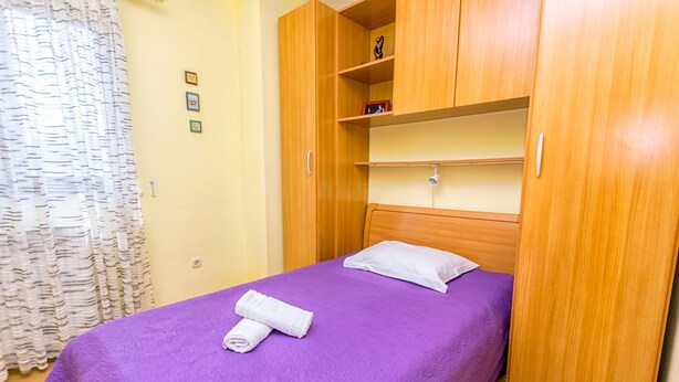 A single bed with a purple bedspread and a white pillow, a wooden wardrobe with shelves, and a curtain with a wavy pattern. - Apartment Leo (Racisce)