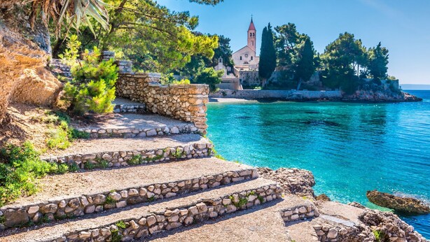 A coastal scene with stone steps leading to a building with a red tower, surrounded by trees and water. - Apartment Anic (Bol)