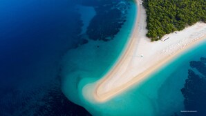 An aerial view of a beach with clear turquoise waters, a sandy shore, and a dense forested area. - Apartment Anic (Bol)