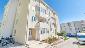 Un immeuble résidentiel à plusieurs étages, doté de balcons, d’un espace de stationnement et d’un petit jardin.