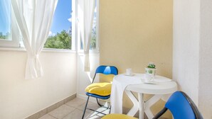 Un petit balcon avec une table et des chaises blanches, une plante en pot et une vue sur les arbres et les montagnes, visible à travers la fenêtre.