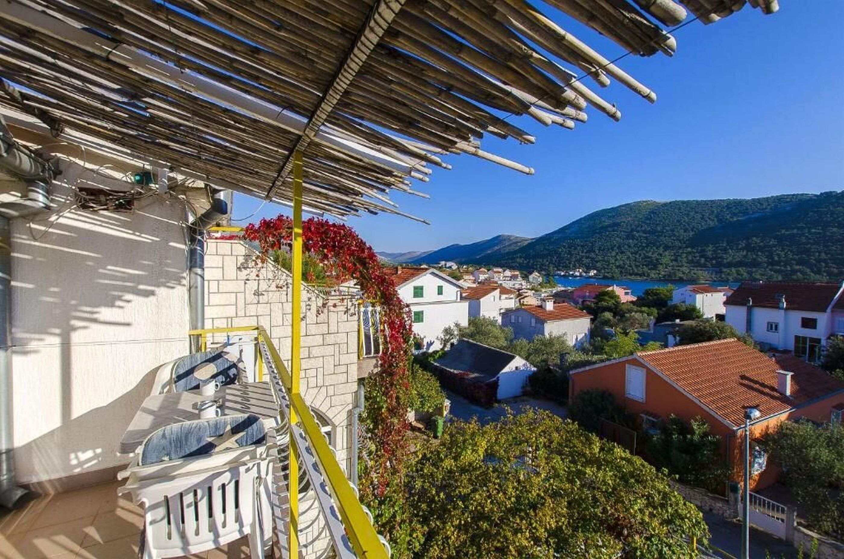 A balcony with a view of a coastal town, a wooden pergola, and a red flowering vine.