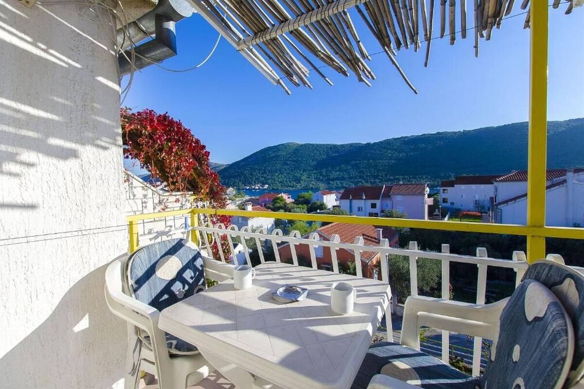 A balcony with a table and chairs, overlooking a coastal town with houses and hills in the background.