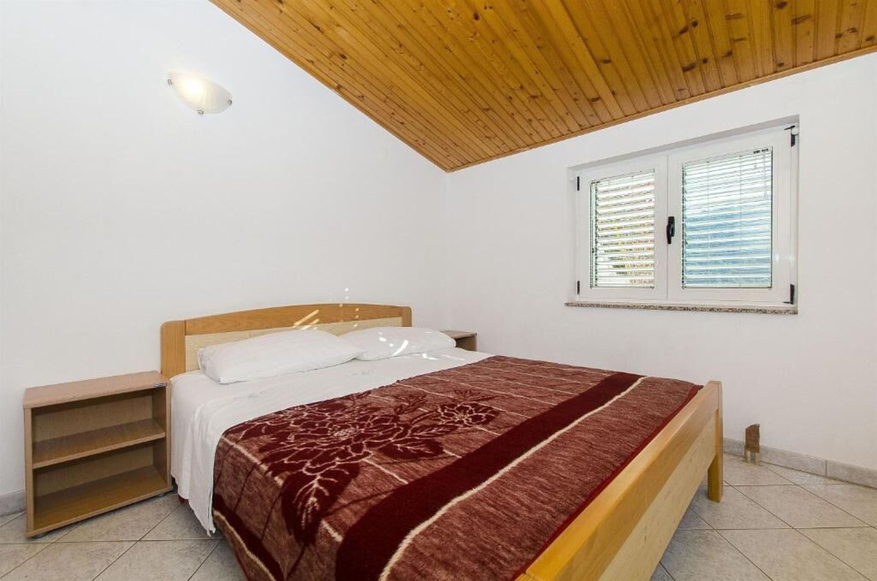 A bedroom with a wooden ceiling, a bed with a red blanket, a nightstand, and a window with blinds.
