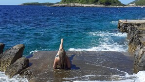 En person står på en klippefyldt kyst med udsigt til havet og et fjerntliggende landskab.