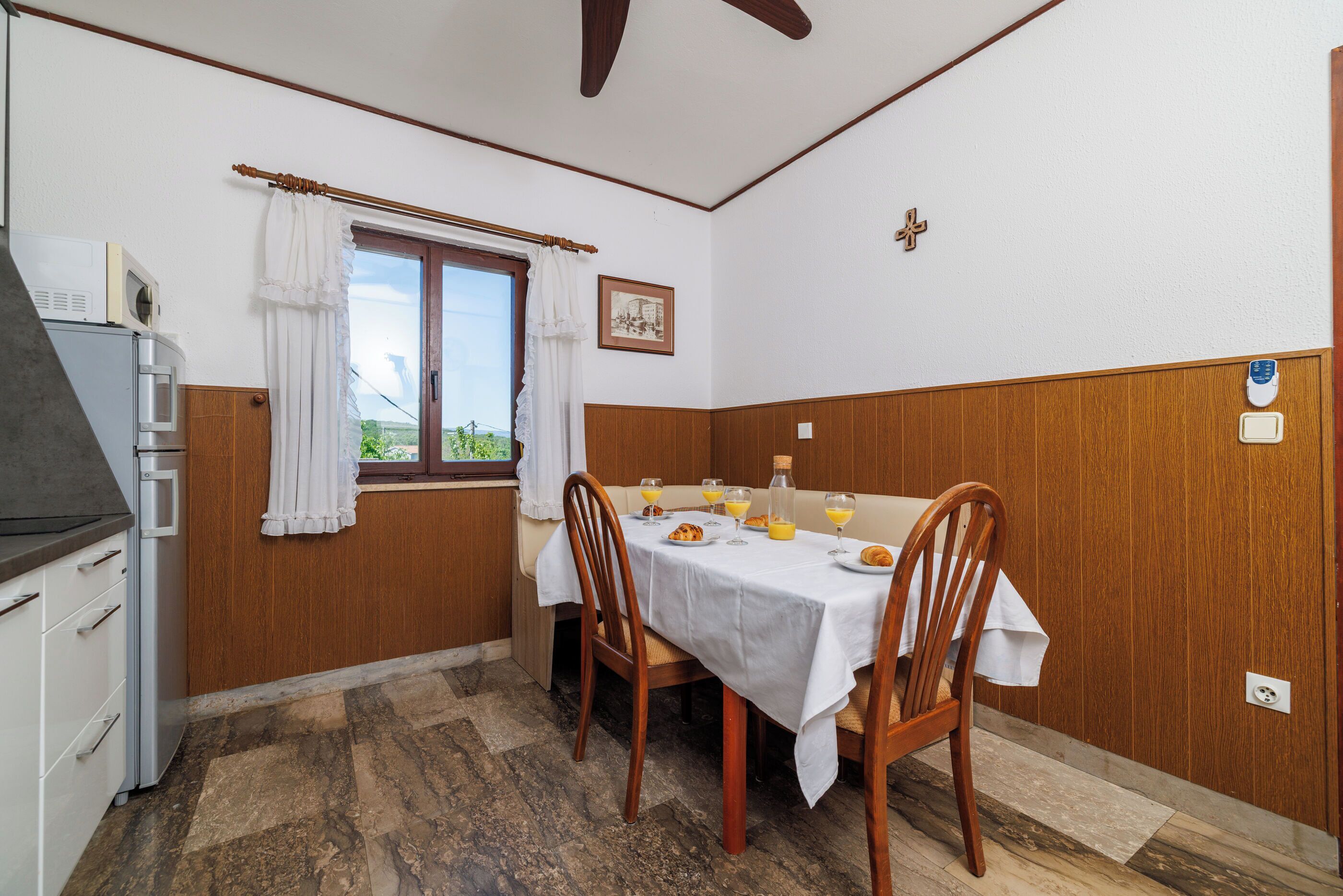 A dining area with a table set for two, a window with curtains, a microwave, and a view of greenery outside.