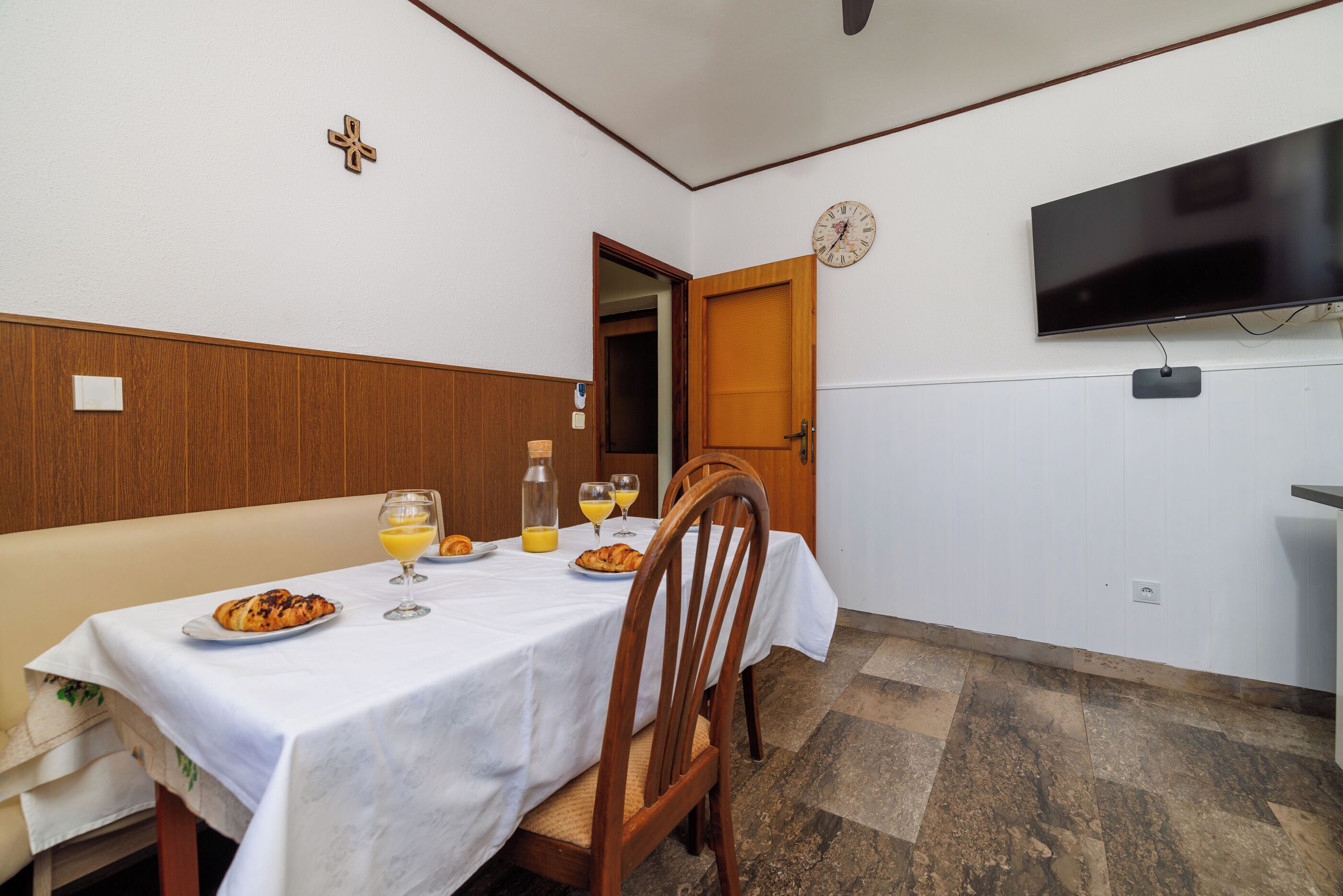 Un coin repas avec une table dressée pour deux personnes, une horloge au mur et un téléviseur fixé au-dessus de la table.