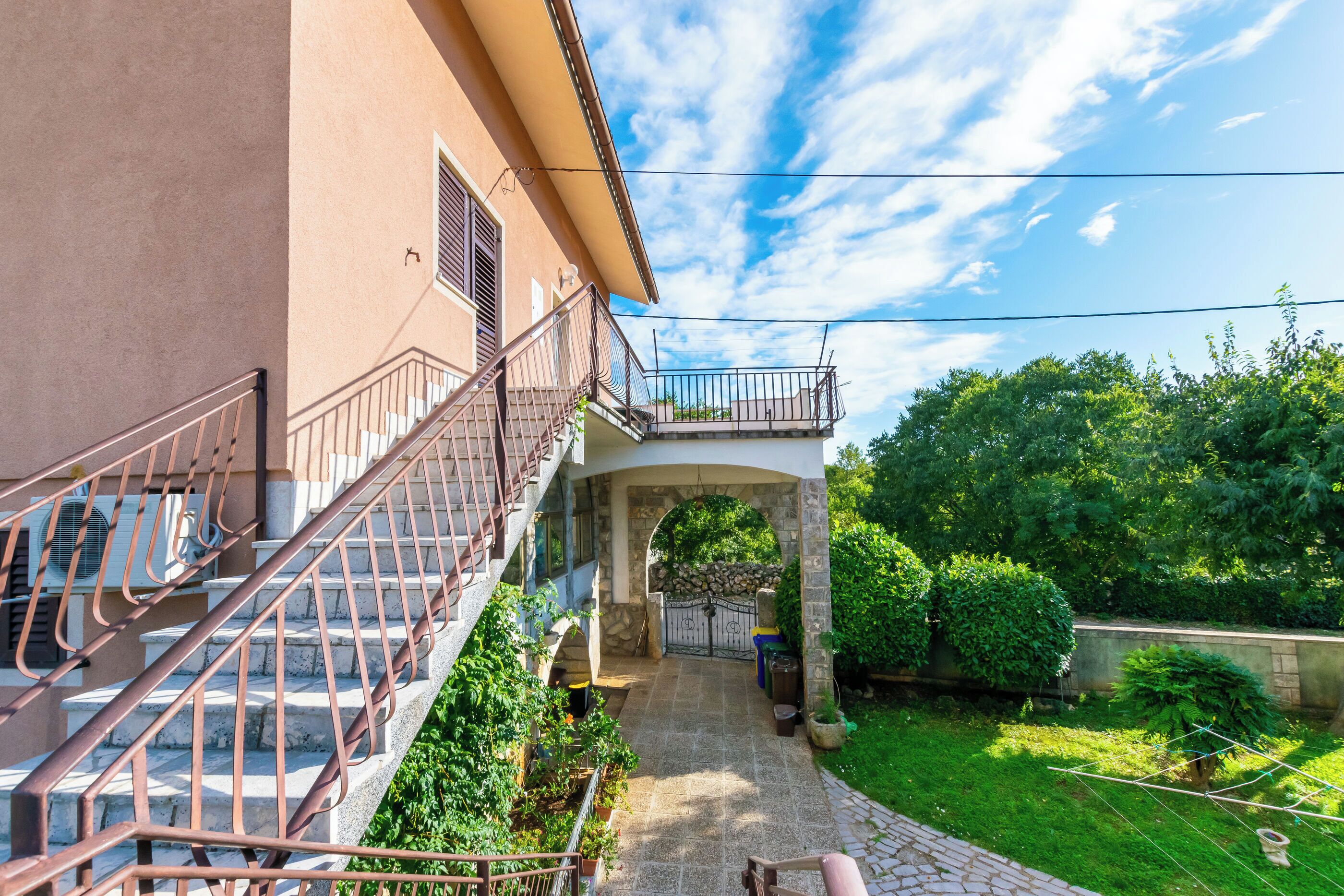 A building with a stone staircase leading to a balcony, surrounded by greenery and a clear sky.