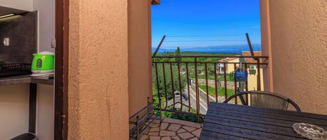 A balcony with a stone floor, a table, and a chair. There is a green basket and a trash bin.
